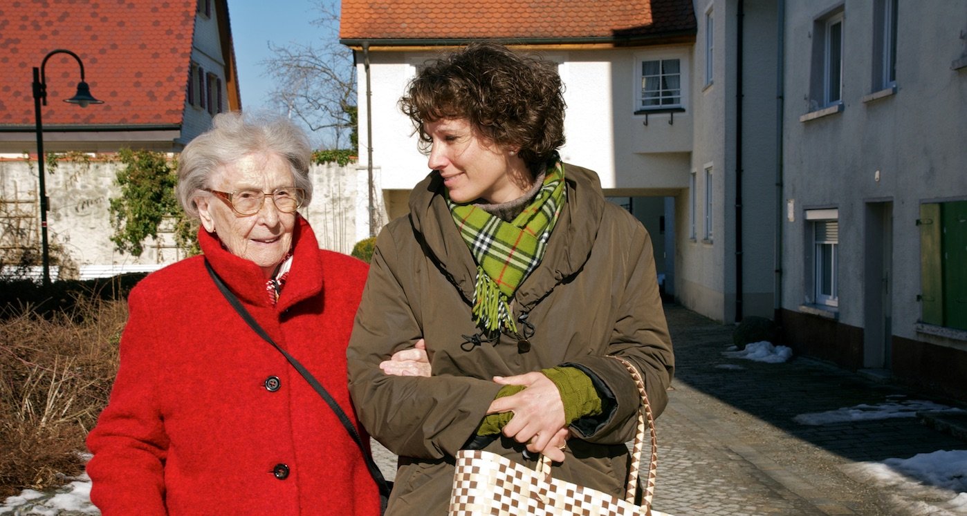 Eine Seniorin im Sessel wird von einer Frau von hinten umarmt