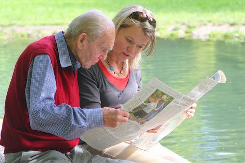 Eine Seniorin im Sessel wird von einer Frau von hinten umarmt