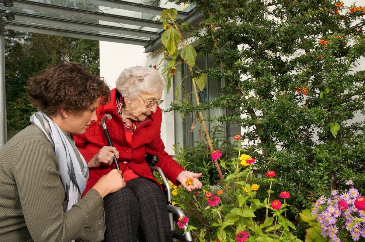 Eine Seniorin im Sessel wird von einer Frau von hinten umarmt