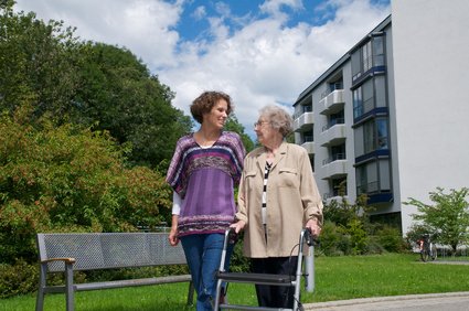 Eine Seniorin im Sessel wird von einer Frau von hinten umarmt