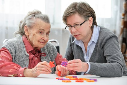 Eine Seniorin im Sessel wird von einer Frau von hinten umarmt