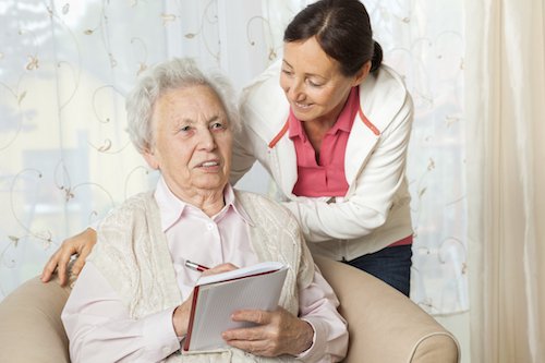 Eine Seniorin im Sessel wird von einer Frau von hinten umarmt
