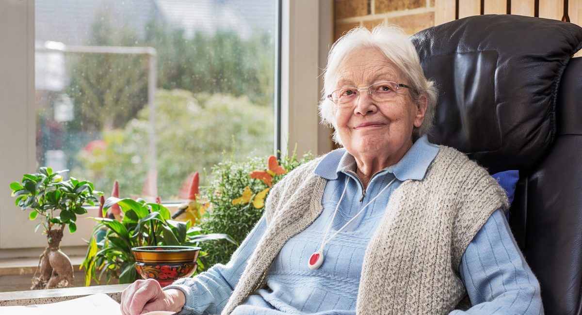 Eine Seniorin im Sessel wird von einer Frau von hinten umarmt