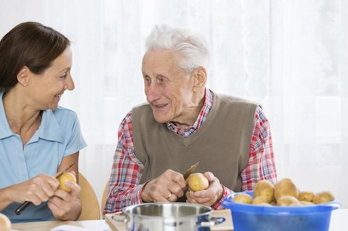 Eine Seniorin im Sessel wird von einer Frau von hinten umarmt