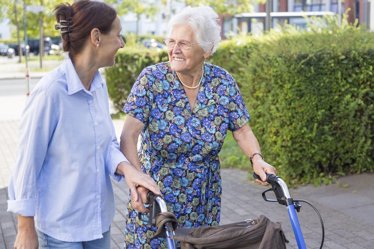 Eine Seniorin mit Rollator wird von einer Frau begleitet